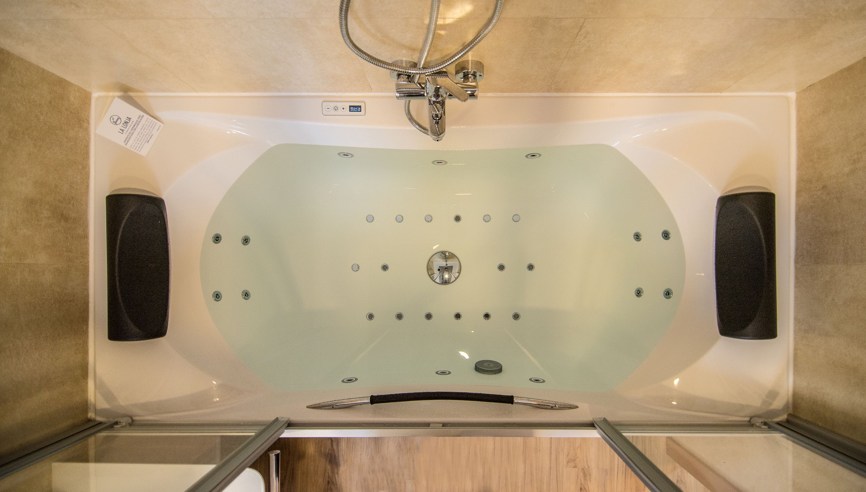Una vasca da bagno ampia e rettangolare con porta in vetro, un soffione doccia sopra e due altoparlanti fissati alla parete.