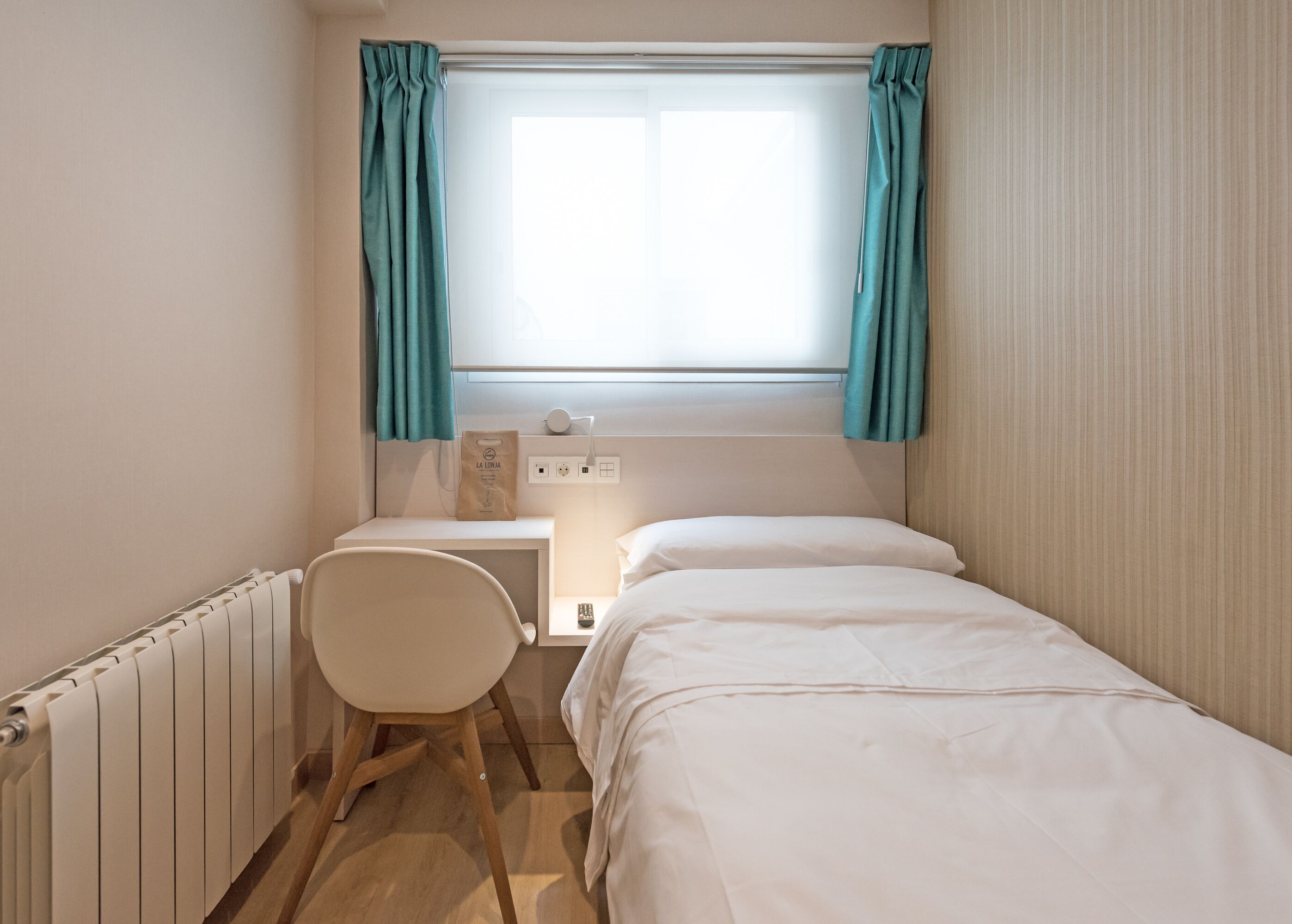 Quarto pequeno e bem iluminado, com uma cama, uma mesa e uma cadeira. Há uma janela com cortinas, um radiador e uma tomada na parede.