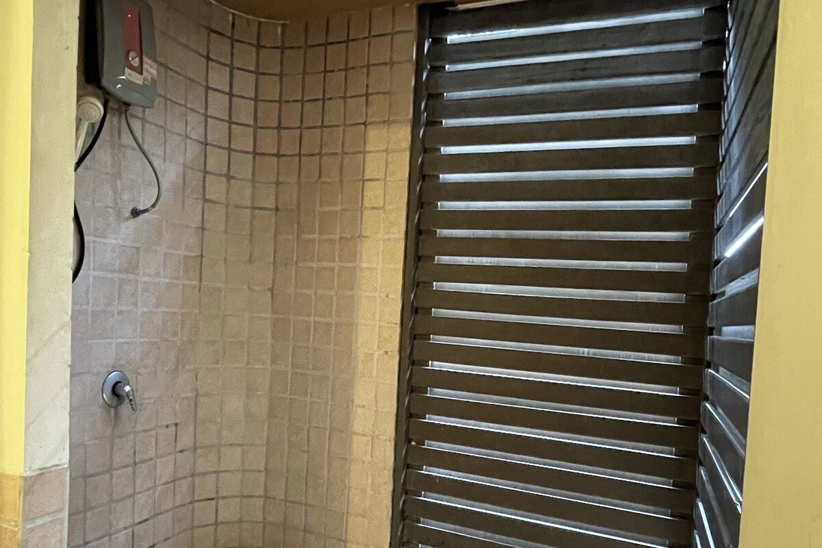 A small, enclosed bathroom with a tiled floor, a toilet, a shower area with a drain, and a window with horizontal blinds.