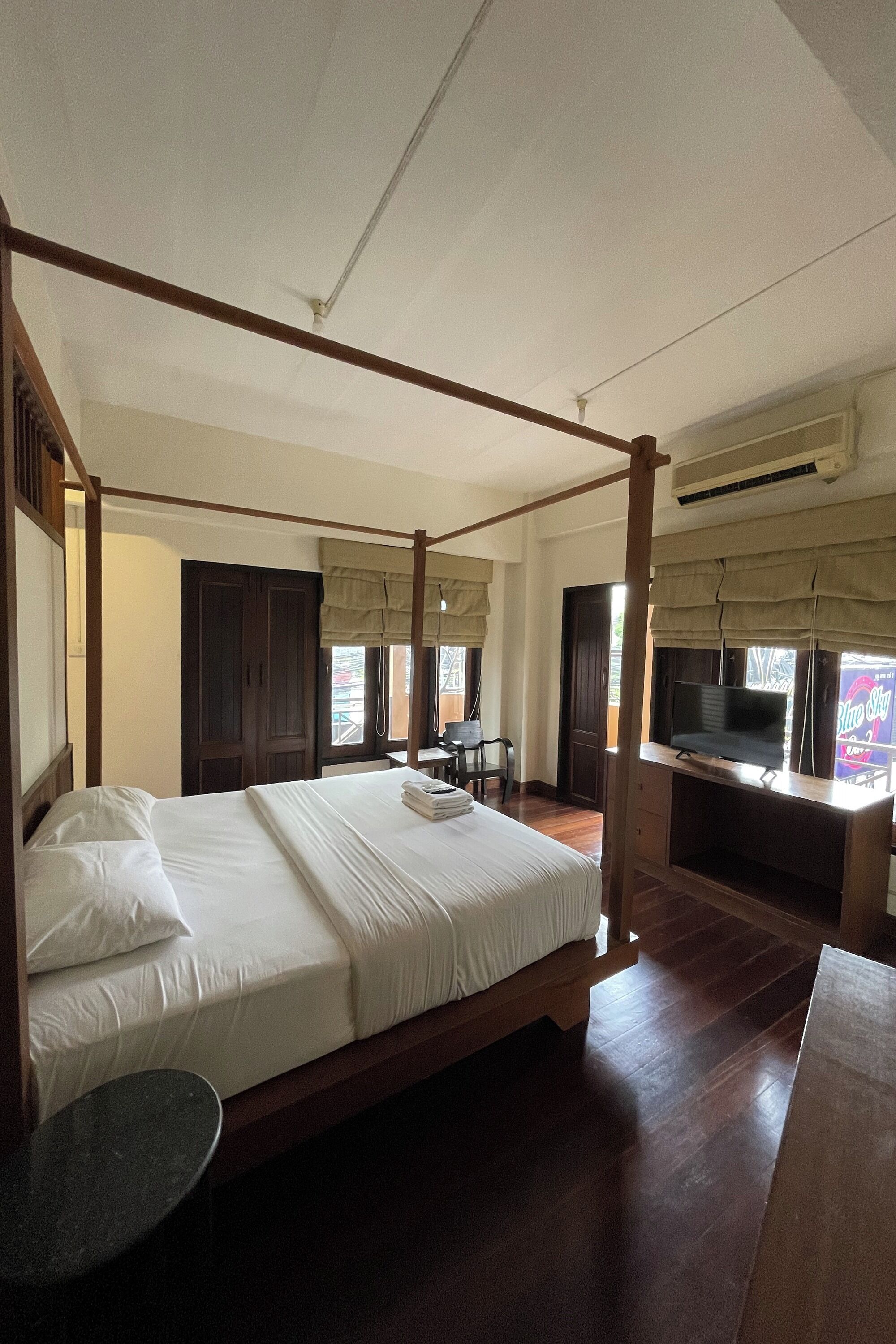 A four-poster bed with a flat-screen TV on a wooden desk, a small round table, and a window with curtains.
