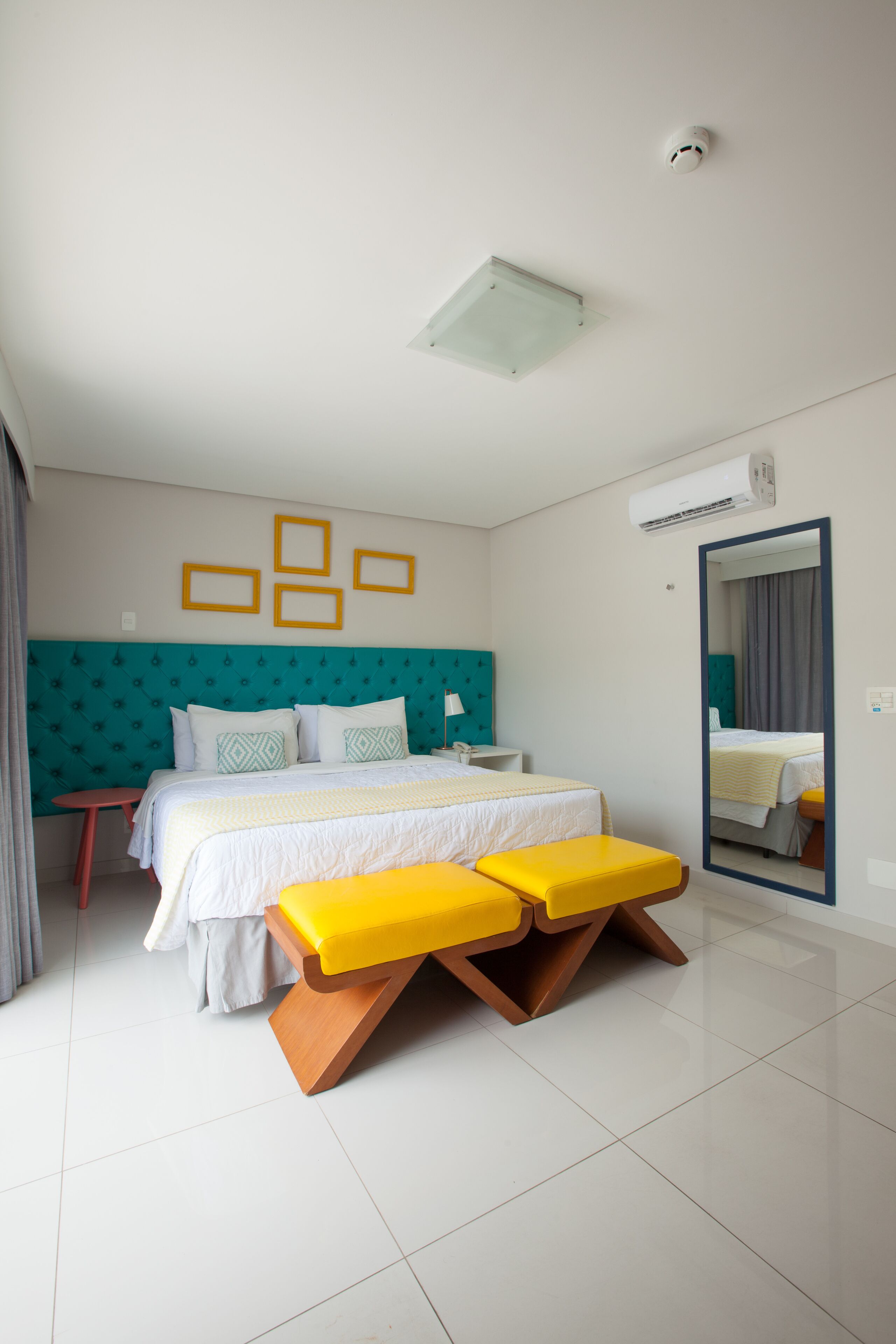 A modern hotel room with a large bed, two bedside tables, a mirror, and a bright yellow bench.