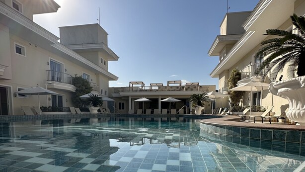 A swimming pool with a tiled floor, surrounded by lounge chairs and umbrellas, with buildings in the background. - Hotel Doral Guarujá (Guarujá)