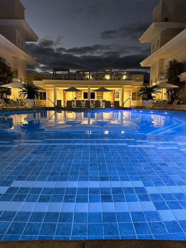 A swimming pool with tiled flooring, surrounded by lounge chairs and umbrellas, with a building in the background.