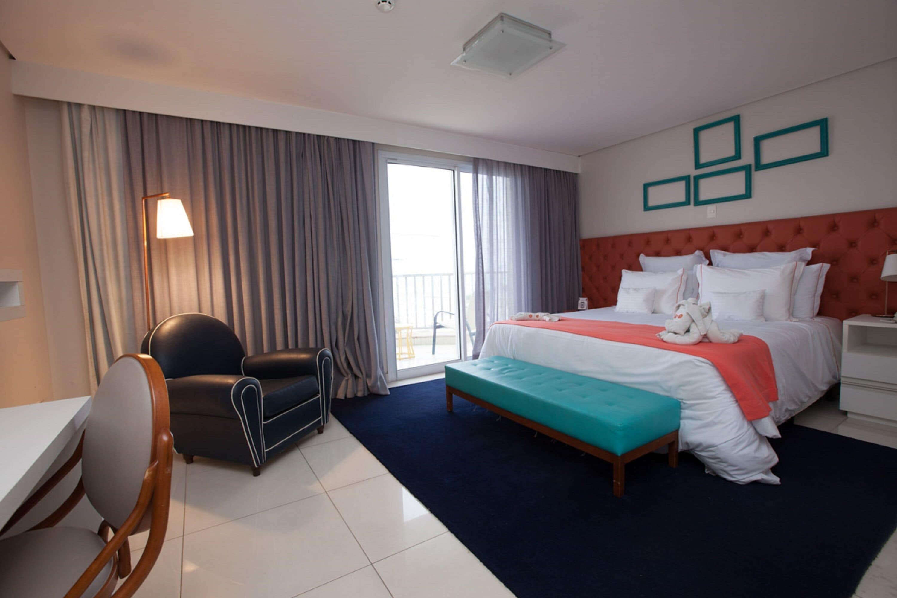 A hotel room with a bed, a desk, a chair, a window with curtains, and a wall with framed pictures.