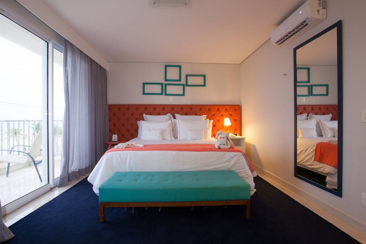 Une chambre d’hôtel avec un grand lit, une banquette, une table de chevet, un miroir et une vue sur l’extérieur.