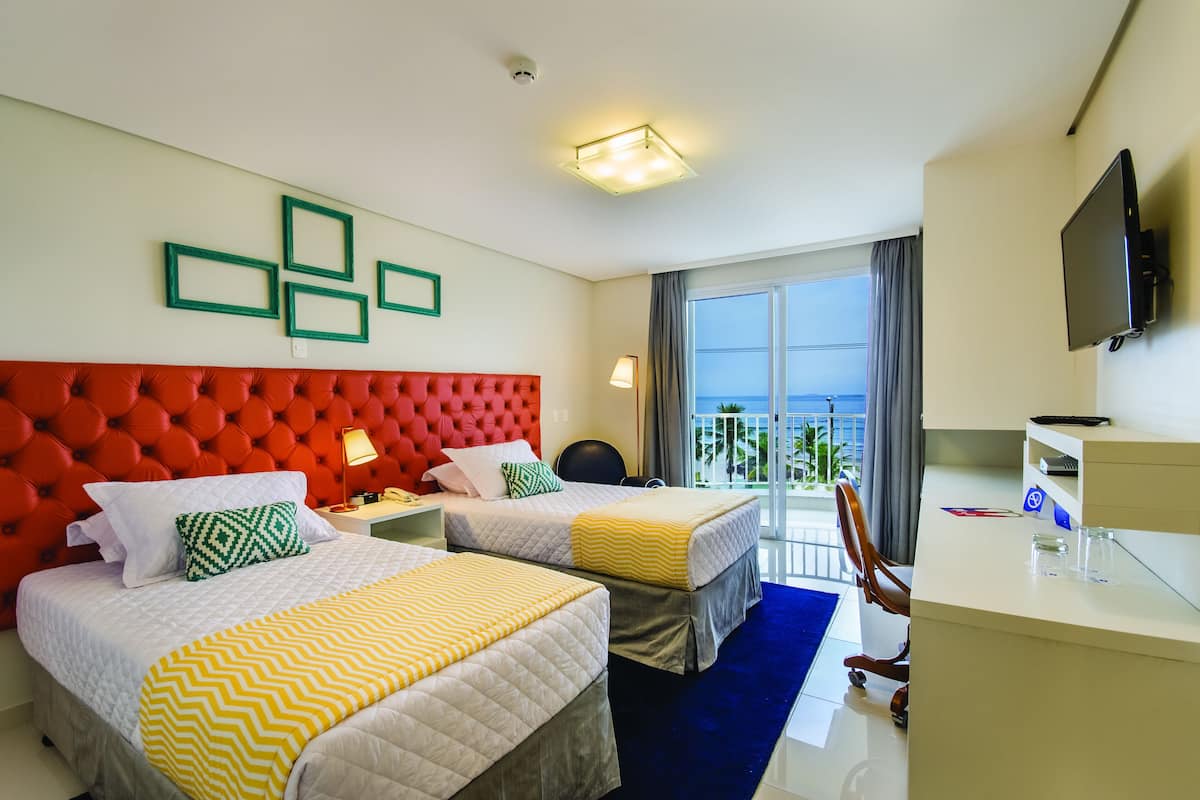 Une chambre d’hôtel avec deux lits, une tête de lit rouge, une télévision, un bureau et un balcon avec vue.