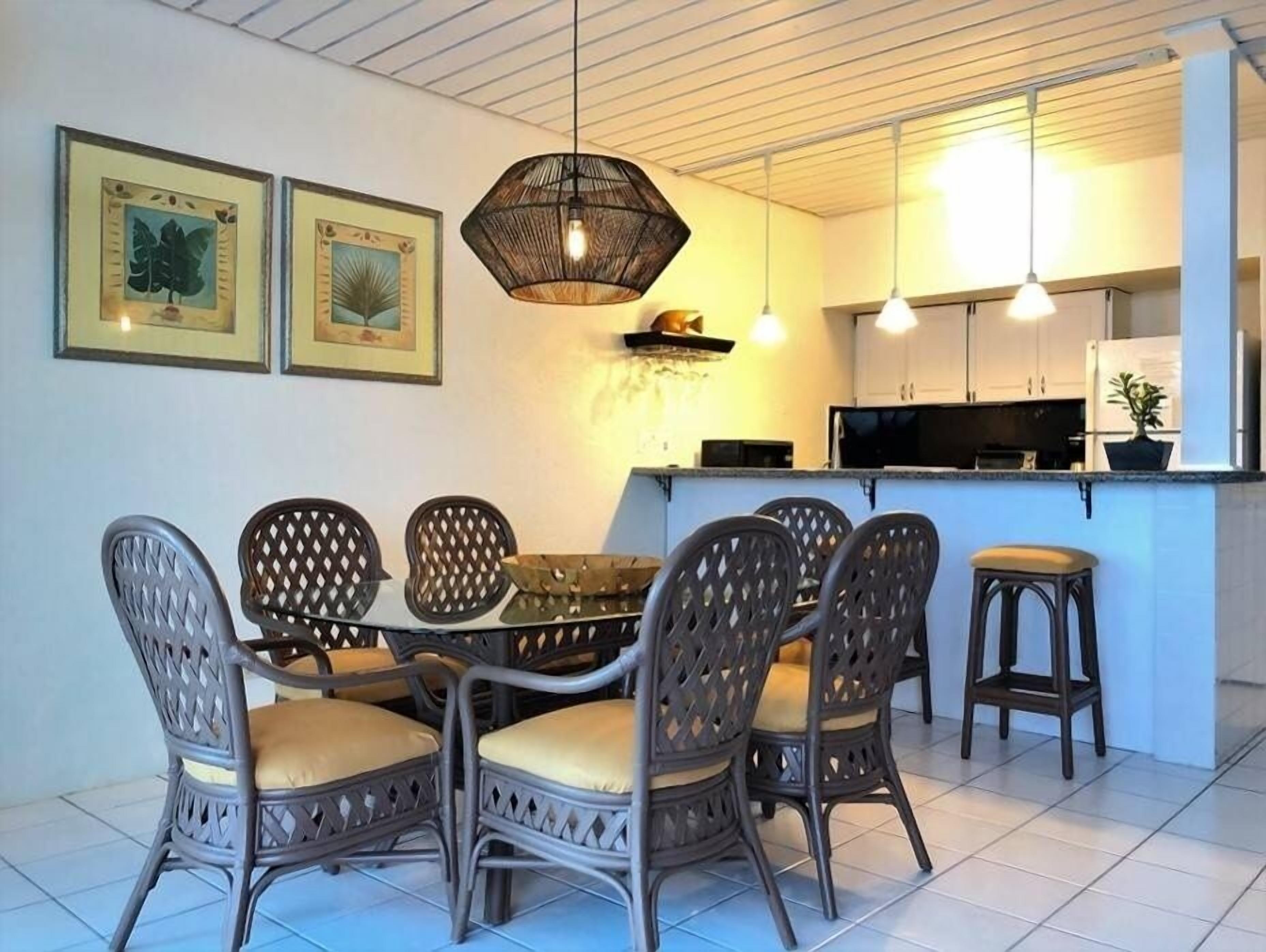 Een eetgedeelte met rieten stoelen en een centrale tafel, een keuken met witte kasten en een magnetron, en twee ingelijste foto’s aan de muur.