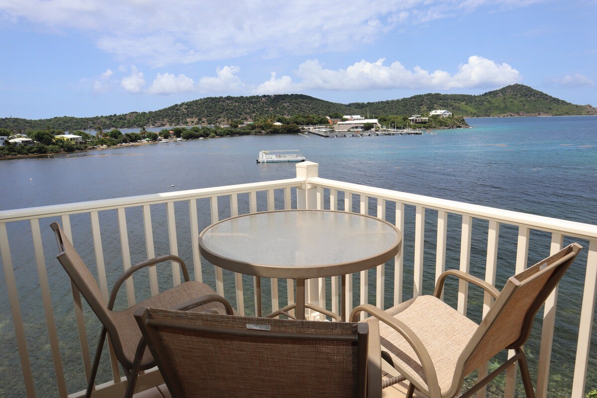 A balcony with a round table and two wicker chairs, overlooking a calm body of water with a small island and buildings in the distance.