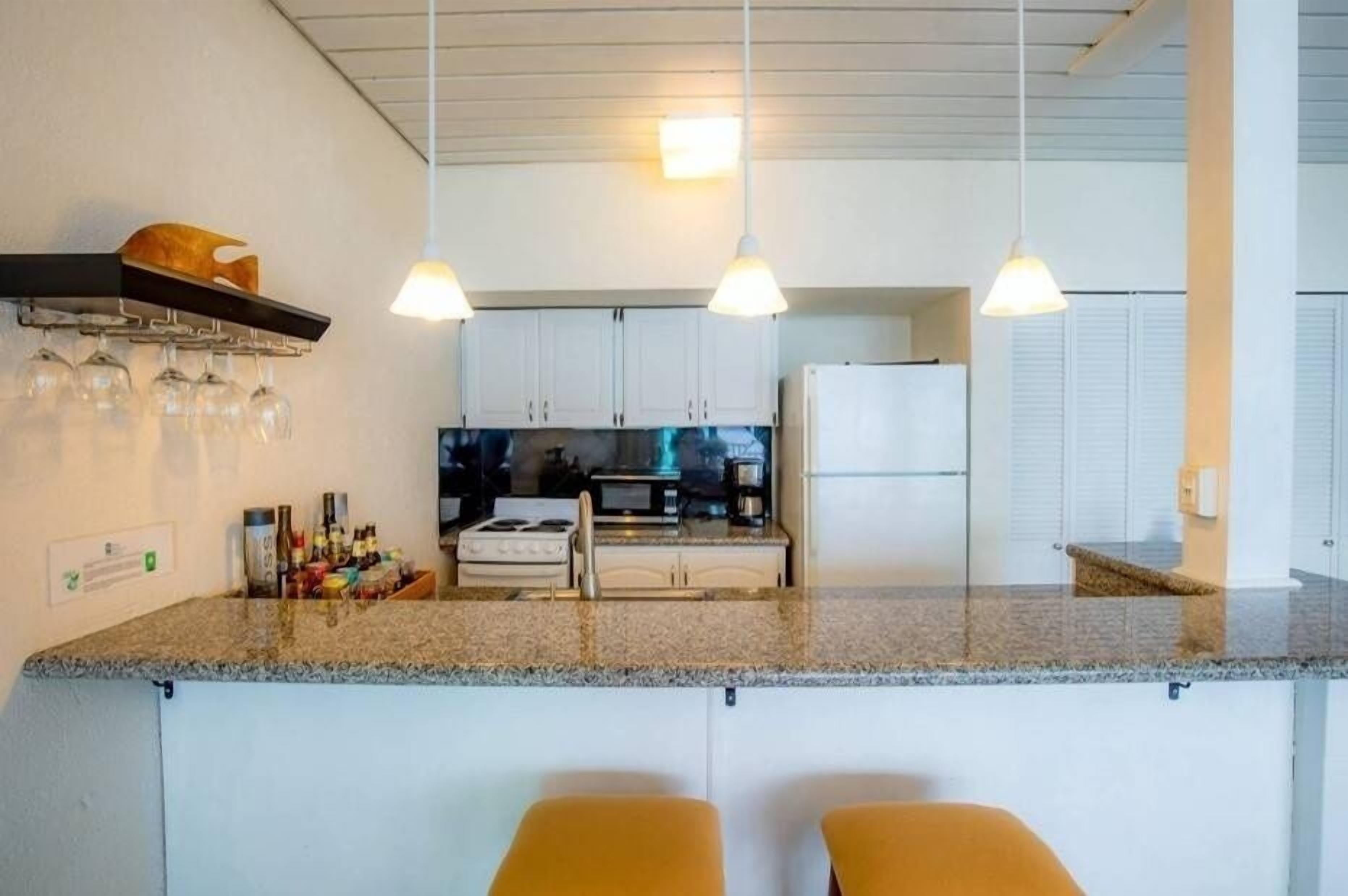 Una cocina con encimeras de granito, gabinetes blancos y una isla central con dos taburetes de bar. Se observan luces colgantes, un microondas y un refrigerador.