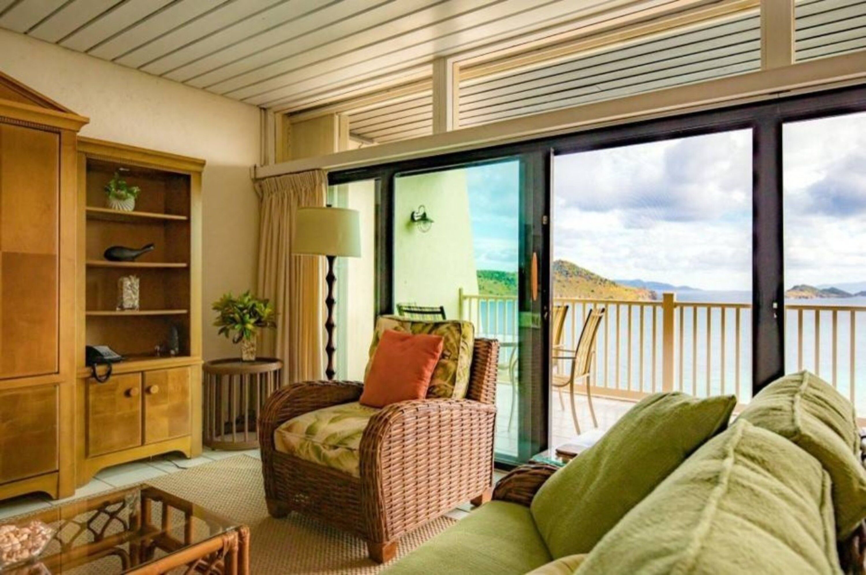 A cozy room with wicker furniture, a glass door leading to a balcony with a view of the sea and mountains.