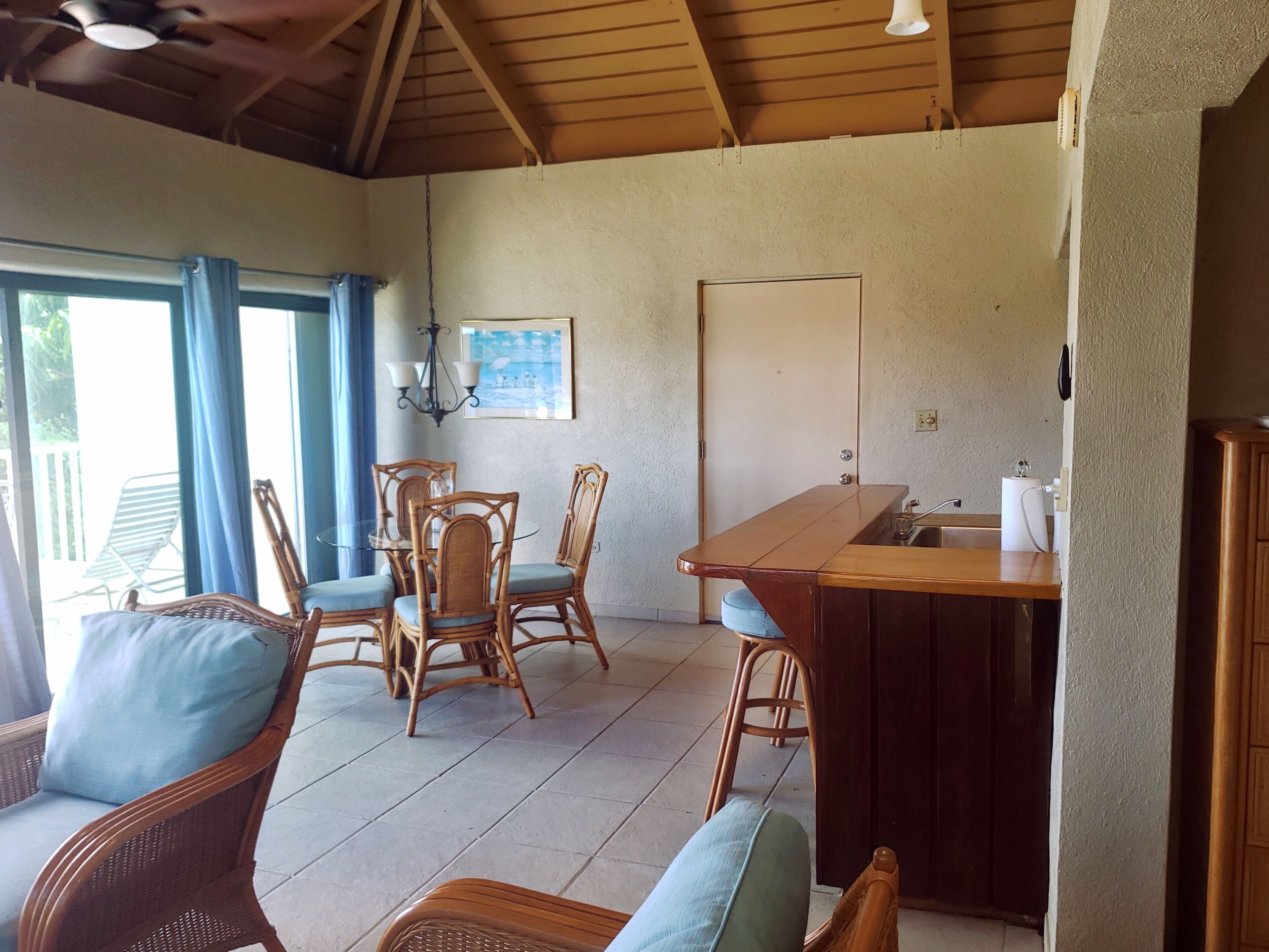 A room with wicker furniture, a glass-top dining table, a kitchen area, and a door.