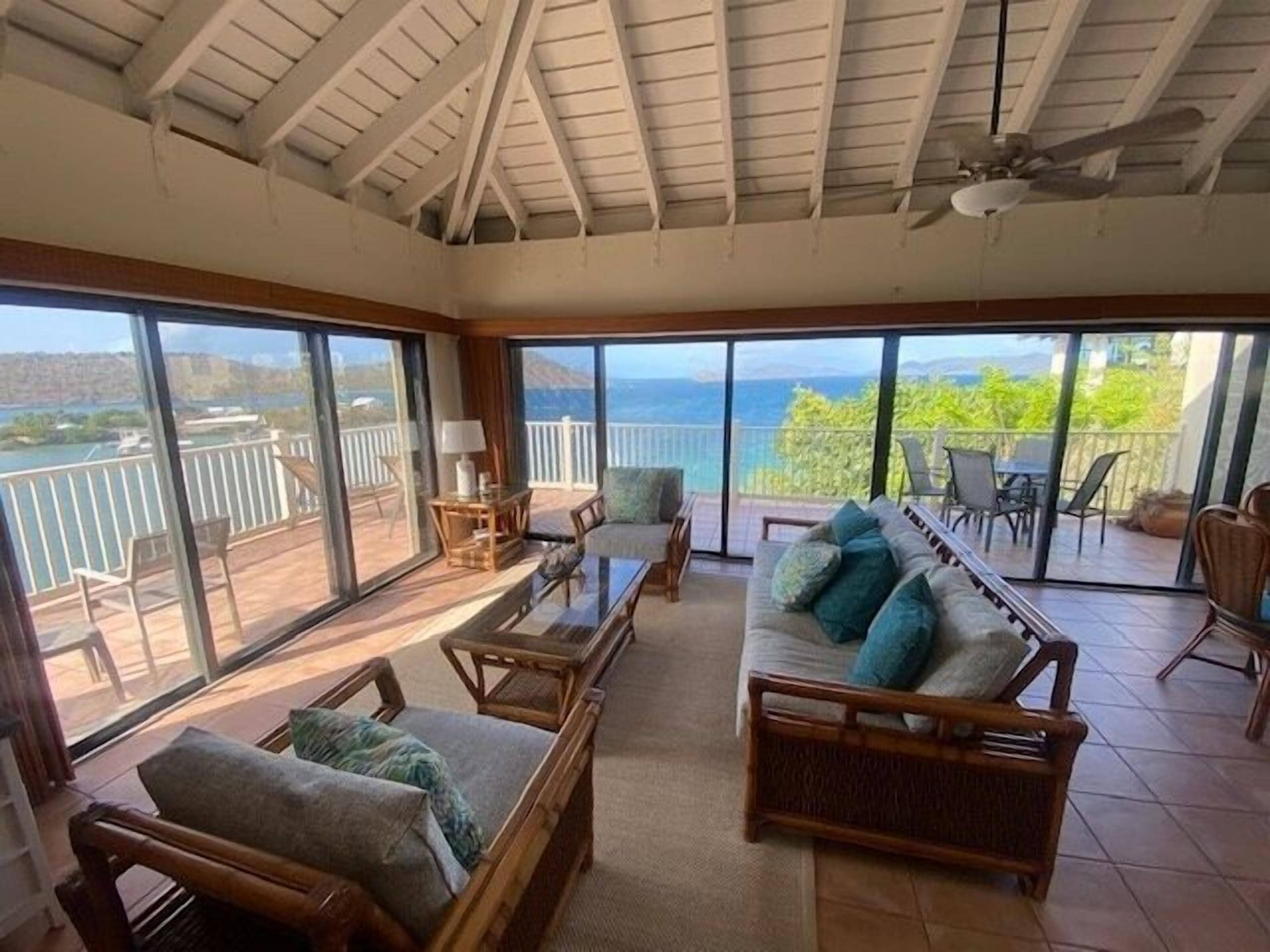 A spacious room with a large sliding glass door leading to a balcony with a view of the sea. There are wicker furniture pieces and a ceiling fan.