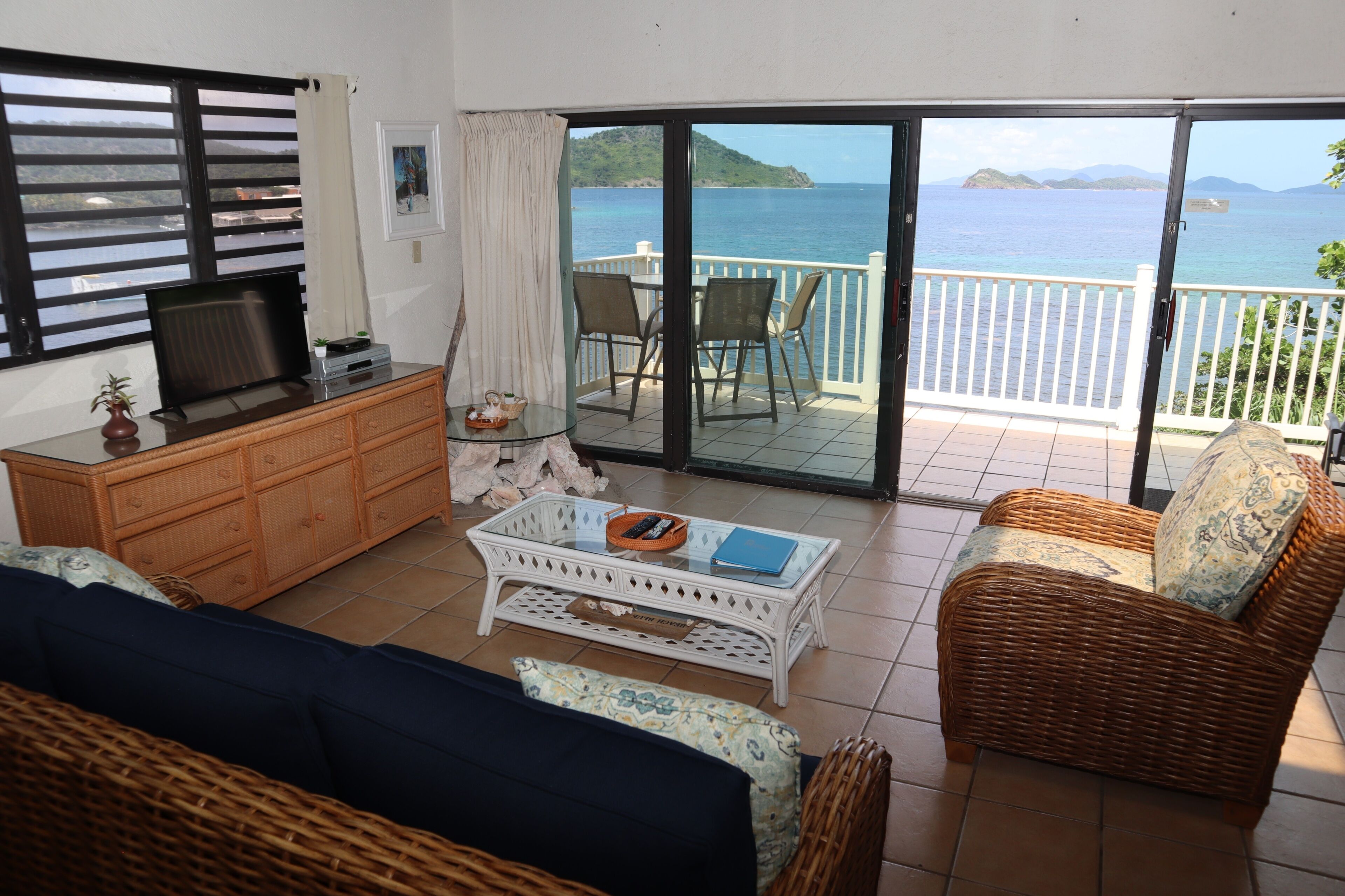 A room with a television, a sofa, a wicker chair, a coffee table, and a balcony with a view of the sea.