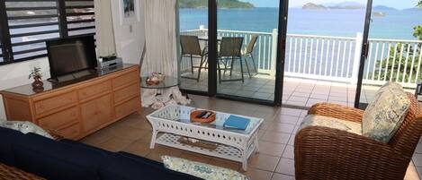 A room with a television, a sofa, a wicker chair, a coffee table, and a balcony with a view of the sea.