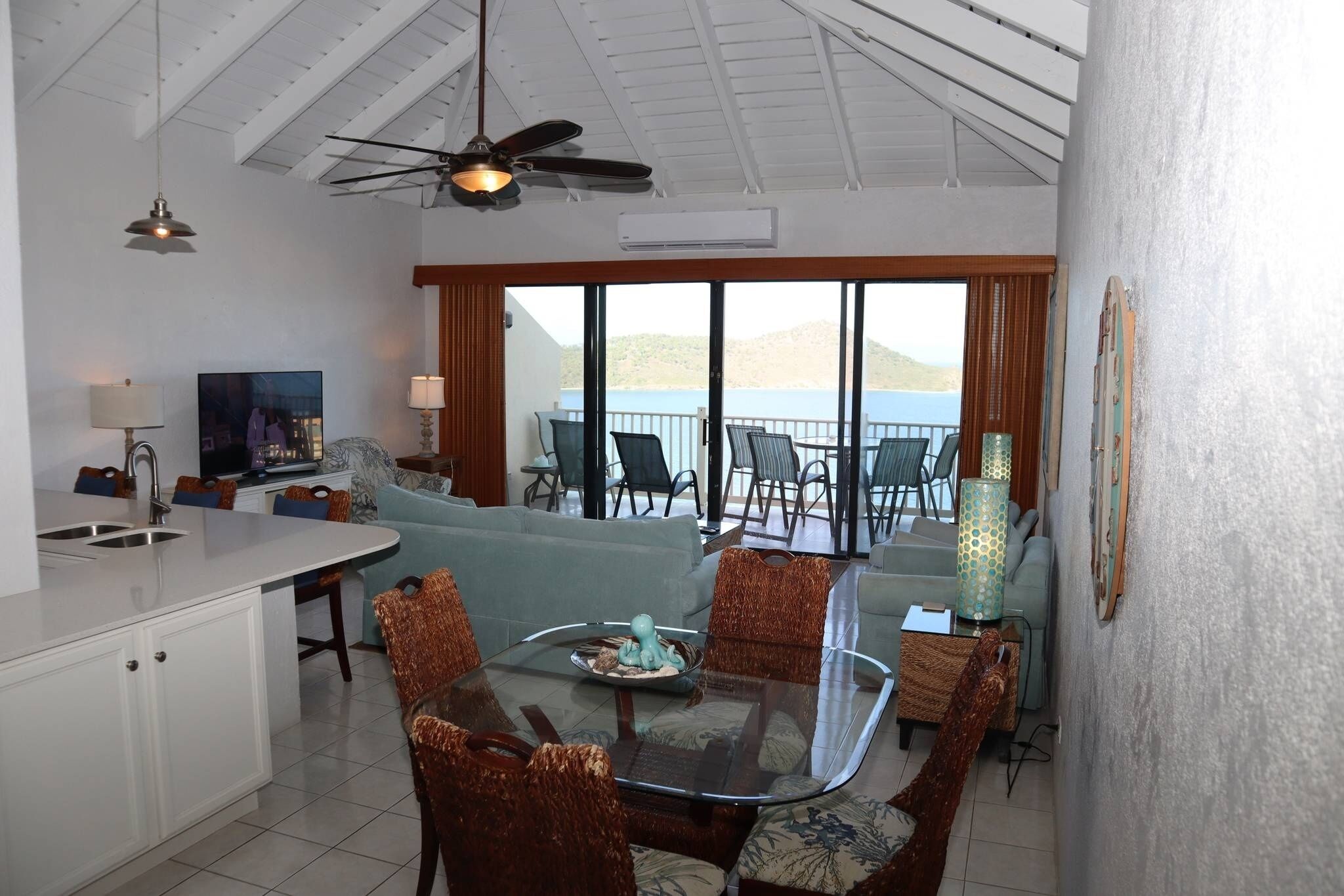 A spacious living area with a glass dining table, comfortable seating, a kitchen area, and a balcony with a view of the sea.