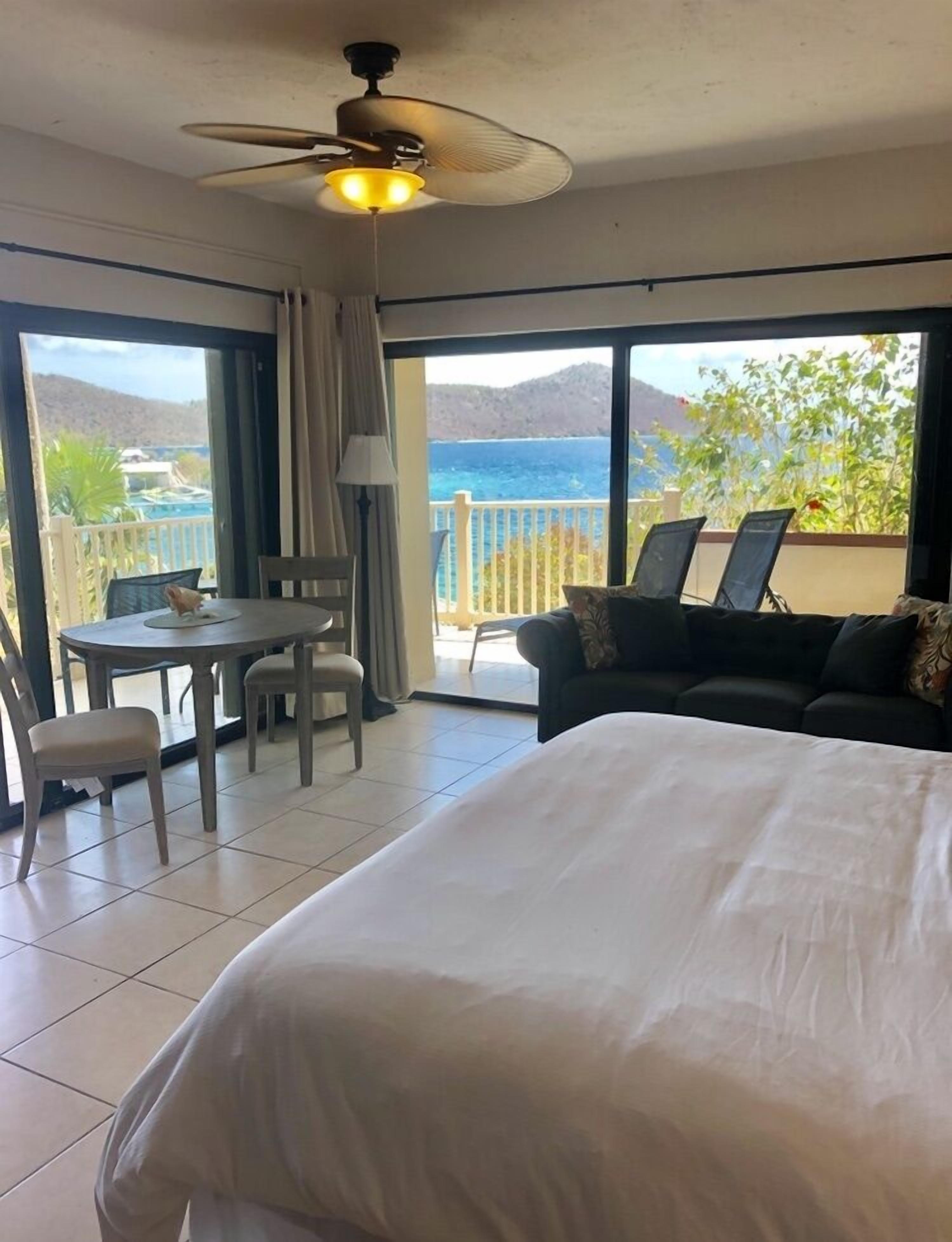 Een hotelkamer met een bed, een eettafel met stoelen, een plafondventilator en een balkon met uitzicht op zee en bergen.