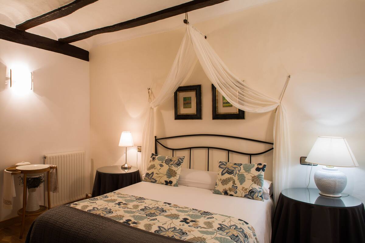 A bedroom with a bed, a bedside table, a nightstand, a lamp, and a window with curtains.