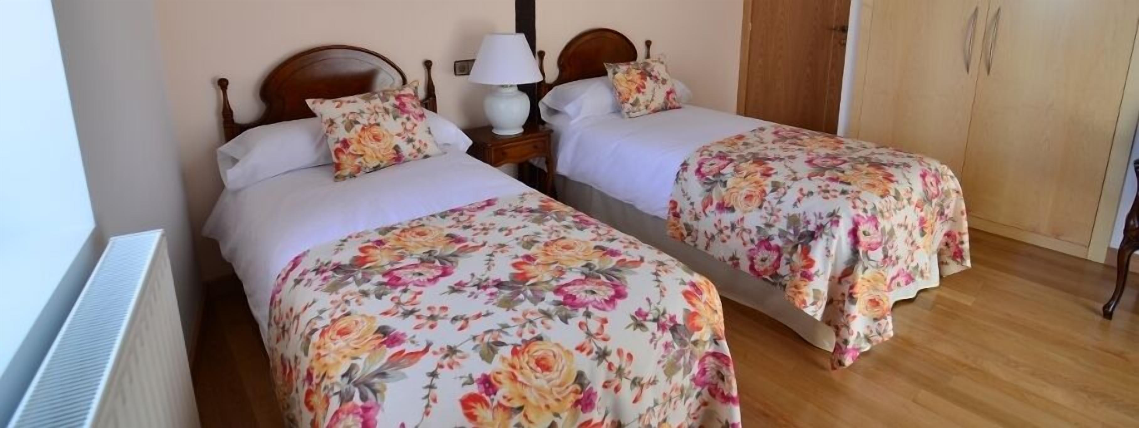 Habitación con dos camas, cada una con ropa de cama floral y un cabecero de madera. Hay una mesita de noche con una lámpara y un armario de madera.