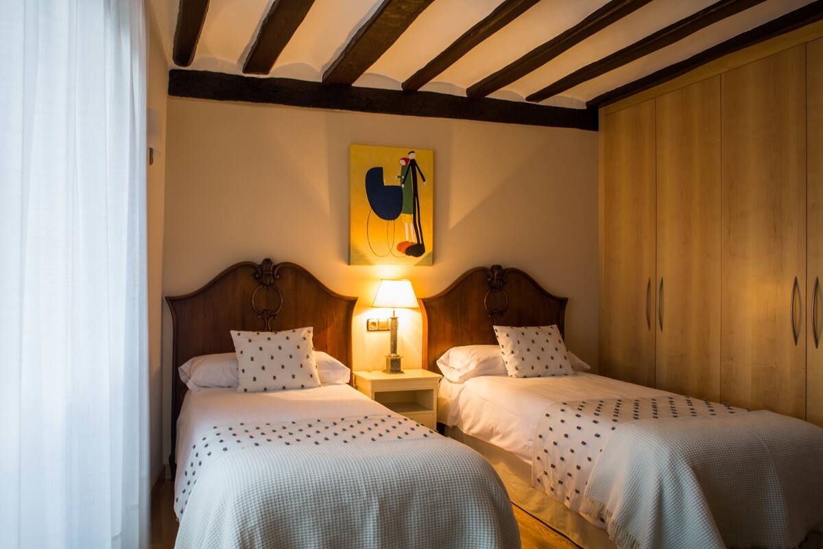 A hotel room with two beds, wooden headboards, a nightstand with a lamp, and a wooden wardrobe.