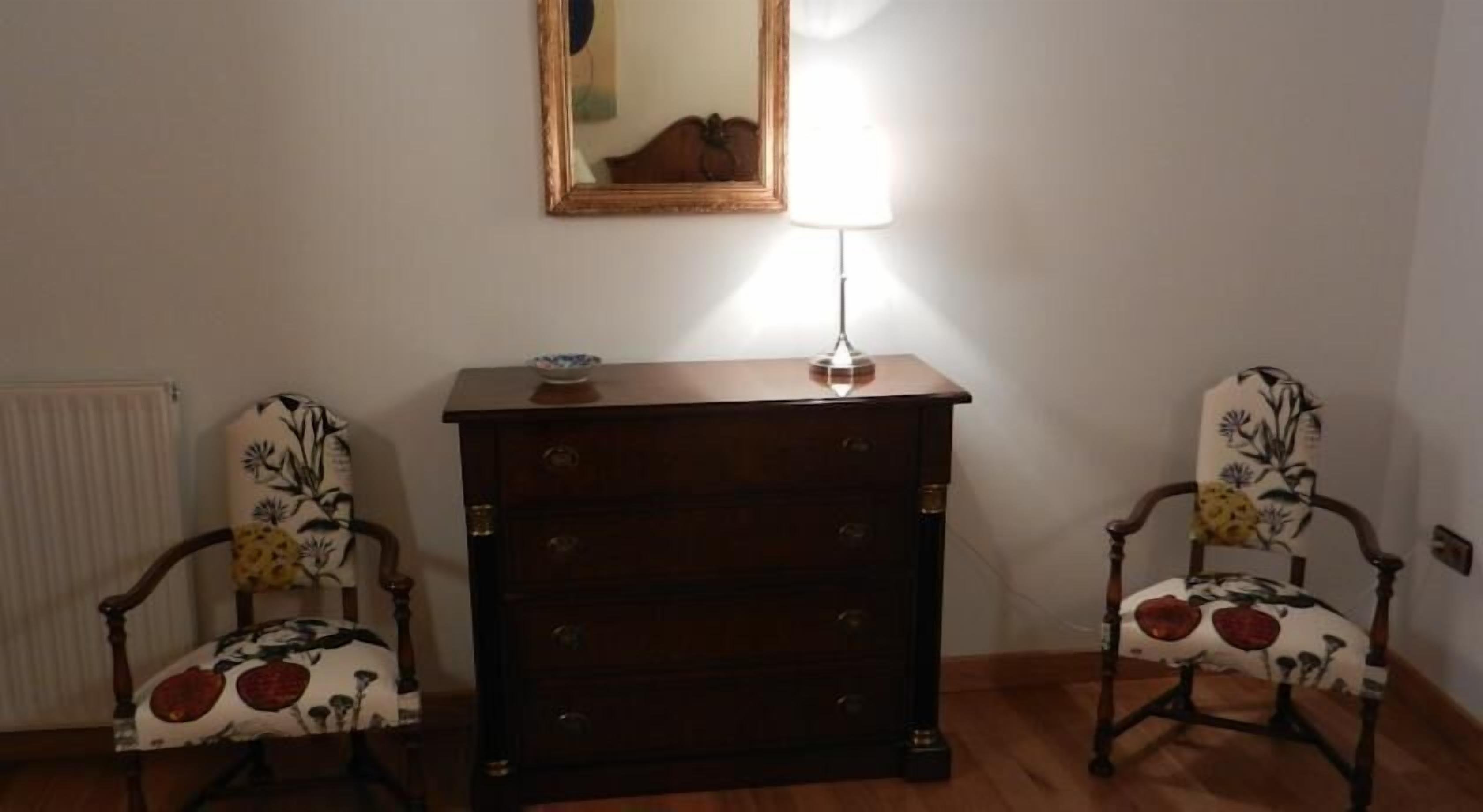 Una habitación con dos sillas de madera tapizadas con motivos florales, un aparador de madera oscura con cajones y un espejo en la pared.
