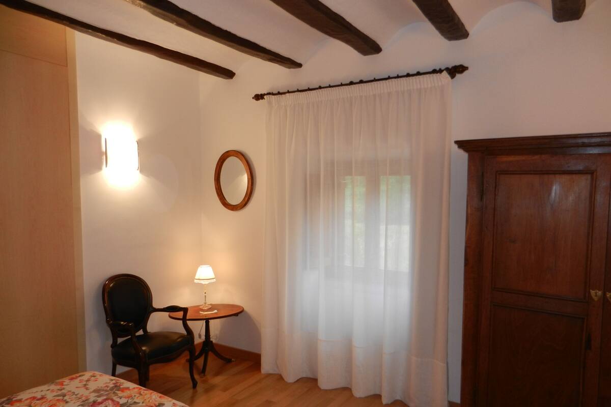 A room with a wooden floor, a bed with a floral bedspread, a small round table with a lamp, a chair, a wooden wardrobe, and a window with sheer curtains.