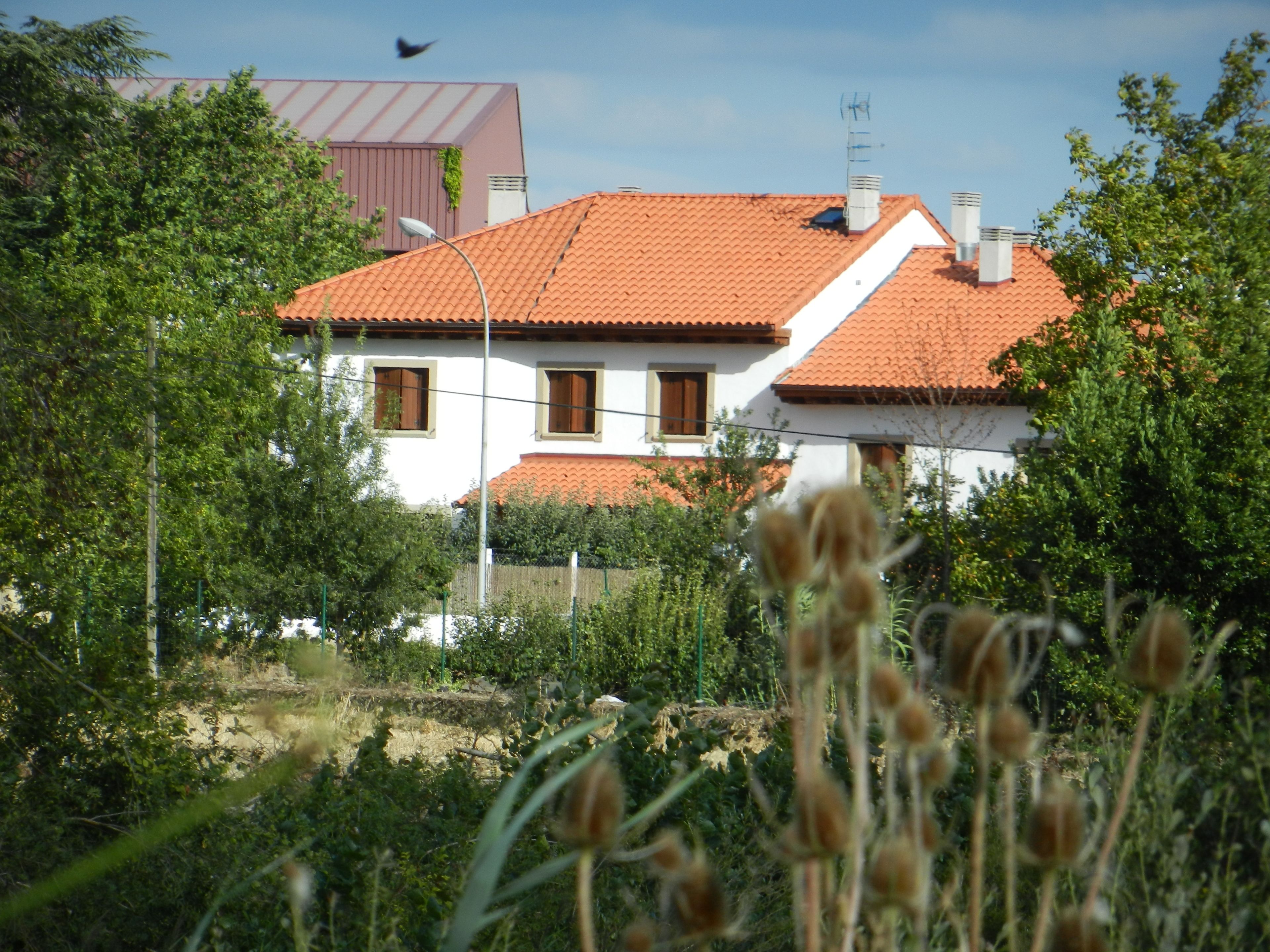 A two-story house with a red-tiled roof, surrounded by greenery and a fence.