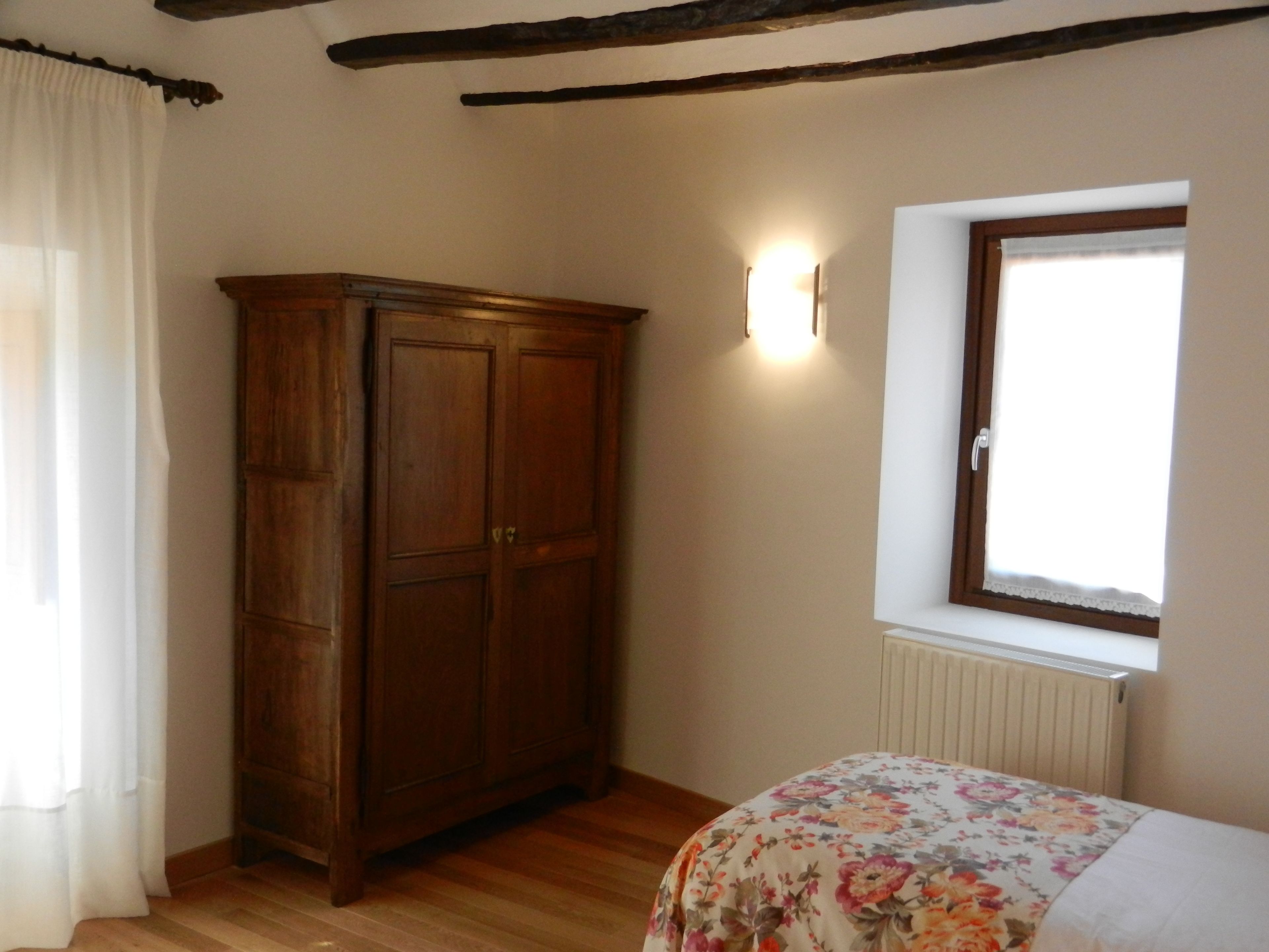 Een slaapkamer met een houten garderobe, een bloemenprint bedspreider, een raam met een witte jaloezie en een wandlamp.