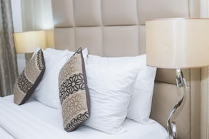 Una cama bien hecha con sábanas blancas y almohadas estampadas, un cabecero color beige y una lámpara de noche.