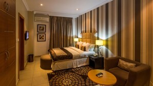 A hotel room with a bed, a sofa, a small table, a TV, and a lamp. - Morning Side Suites (Lagos)