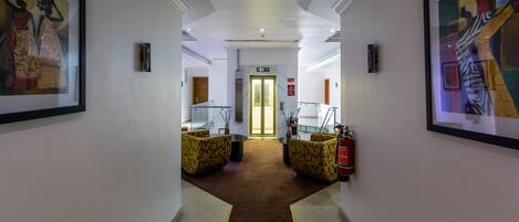 A hotel hallway with framed artwork on the walls, a carpeted floor, and a fire extinguisher on the right.