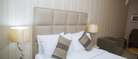 En snyggt bäddad säng med mönstrade kuddar, två sänglampor och en stol i ett hotellrum med randigt tapet.