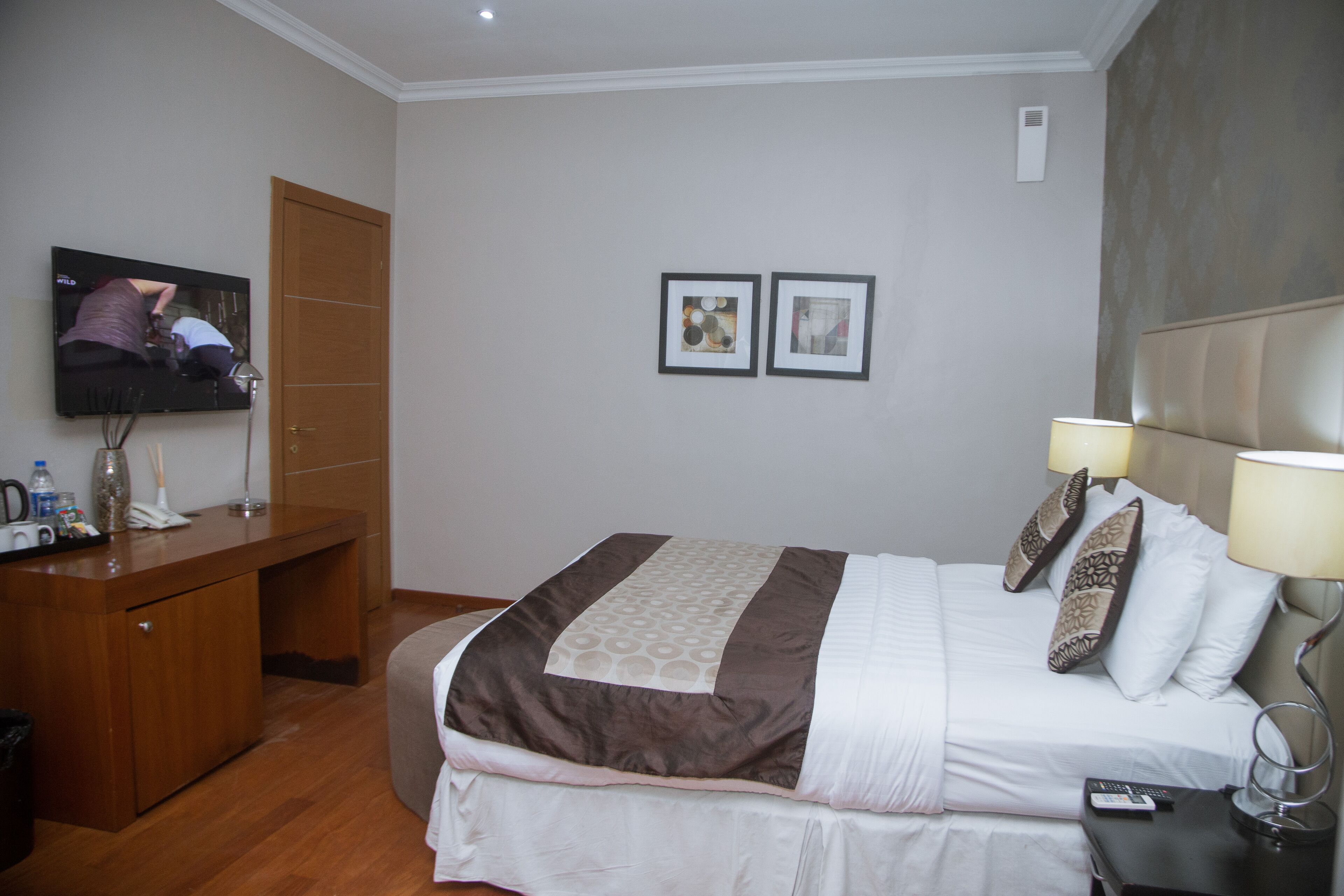 Quarto de hotel com cama, mesa de cabeceira, luminária, televisão e escrivaninha com cadeira.