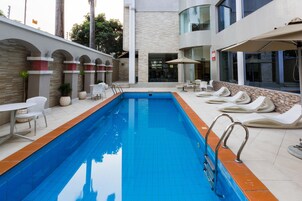 Una piscina con sillones de descanso y mesas, rodeada por un edificio de fachada de piedra y ventanas de vidrio.