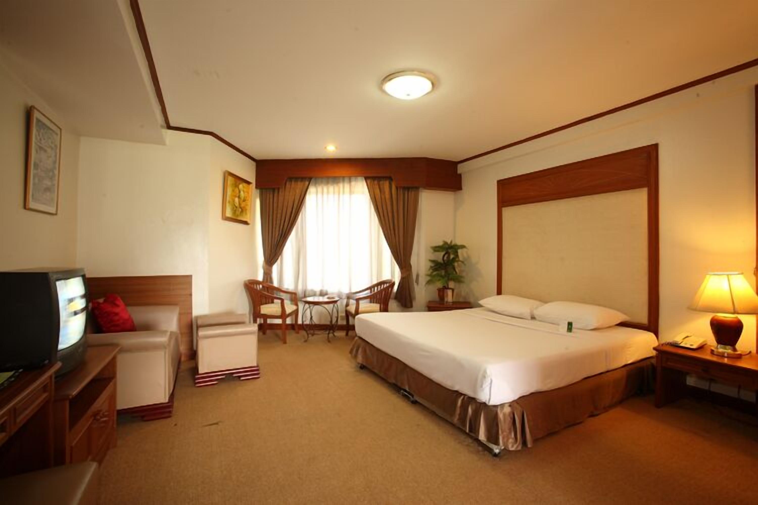 A hotel room with a bed, a desk with a television, a chair, a small table, and a lamp.