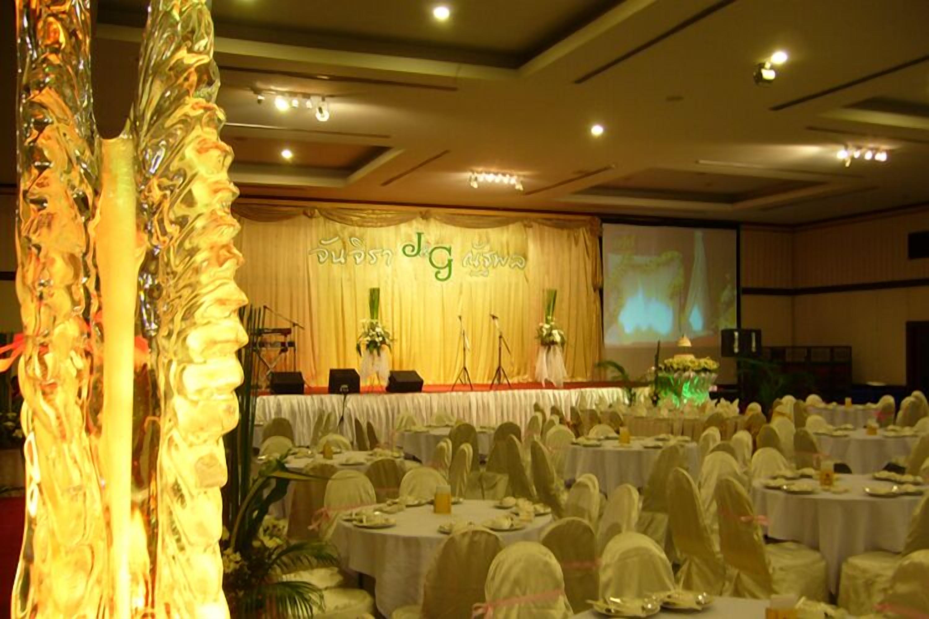Un salón de banquetes con mesas redondas cubiertas con manteles blancos y sillas, un escenario con fondo y una pantalla grande.