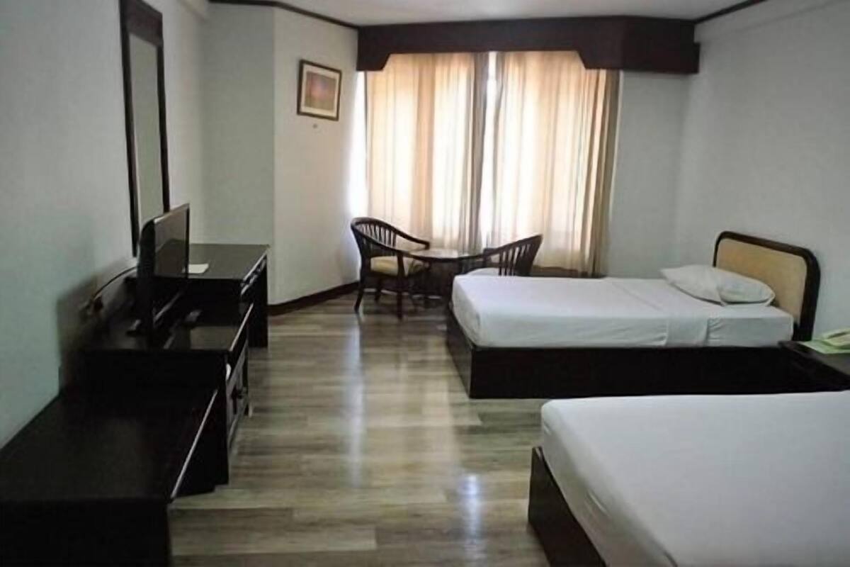A hotel room with two single beds, a desk with a chair, a small table, and a window with curtains.