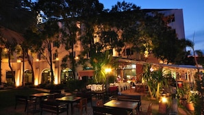 An outdoor dining area with tables and chairs, surrounded by trees and a building with illuminated windows. - Royal Diamond Hotel (Phetchaburi)