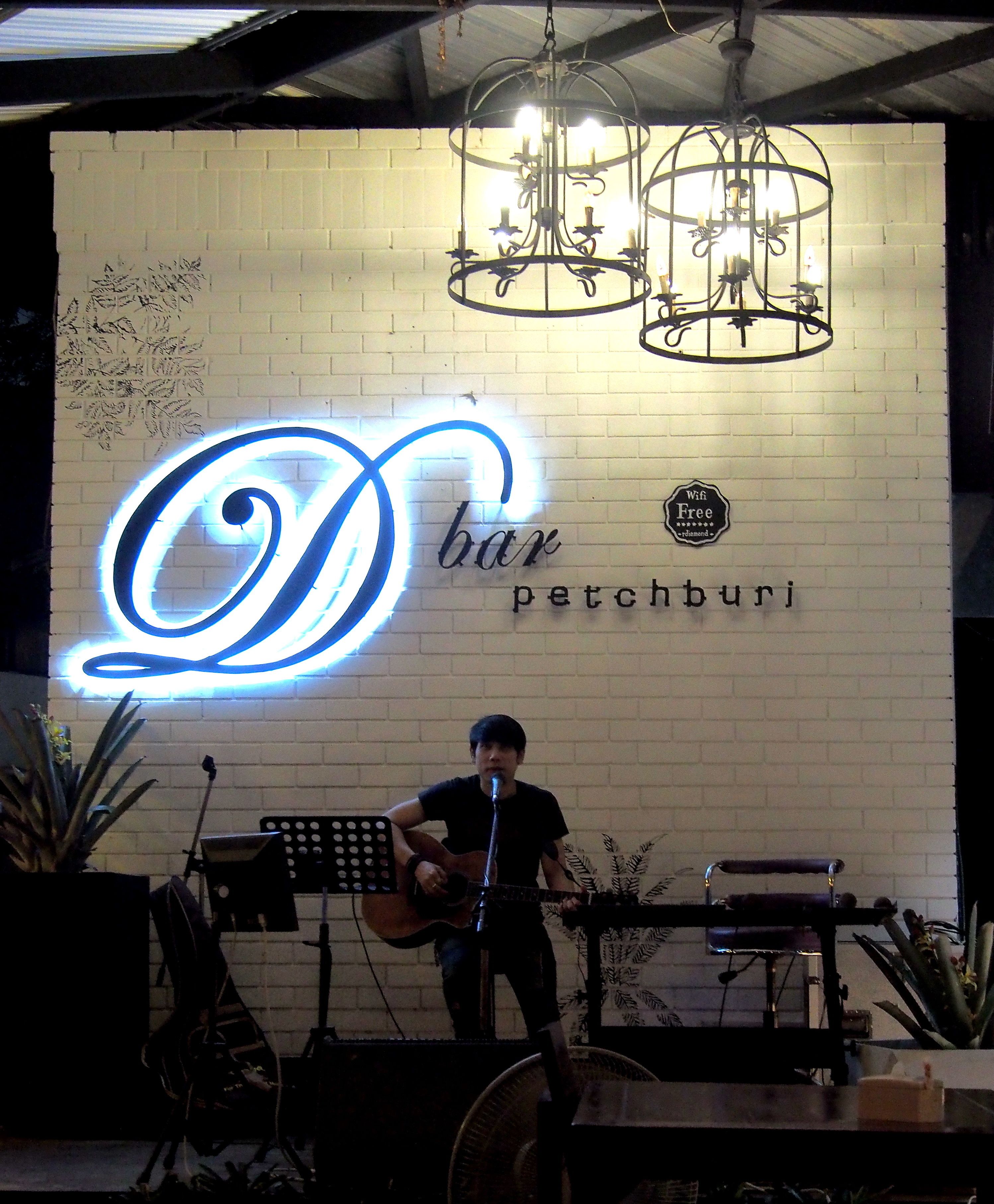 En musiker uppträder på scenen med en neonreklamskylt som visar texten ”D bar petchburi” och en Wi-Fi-symbol.