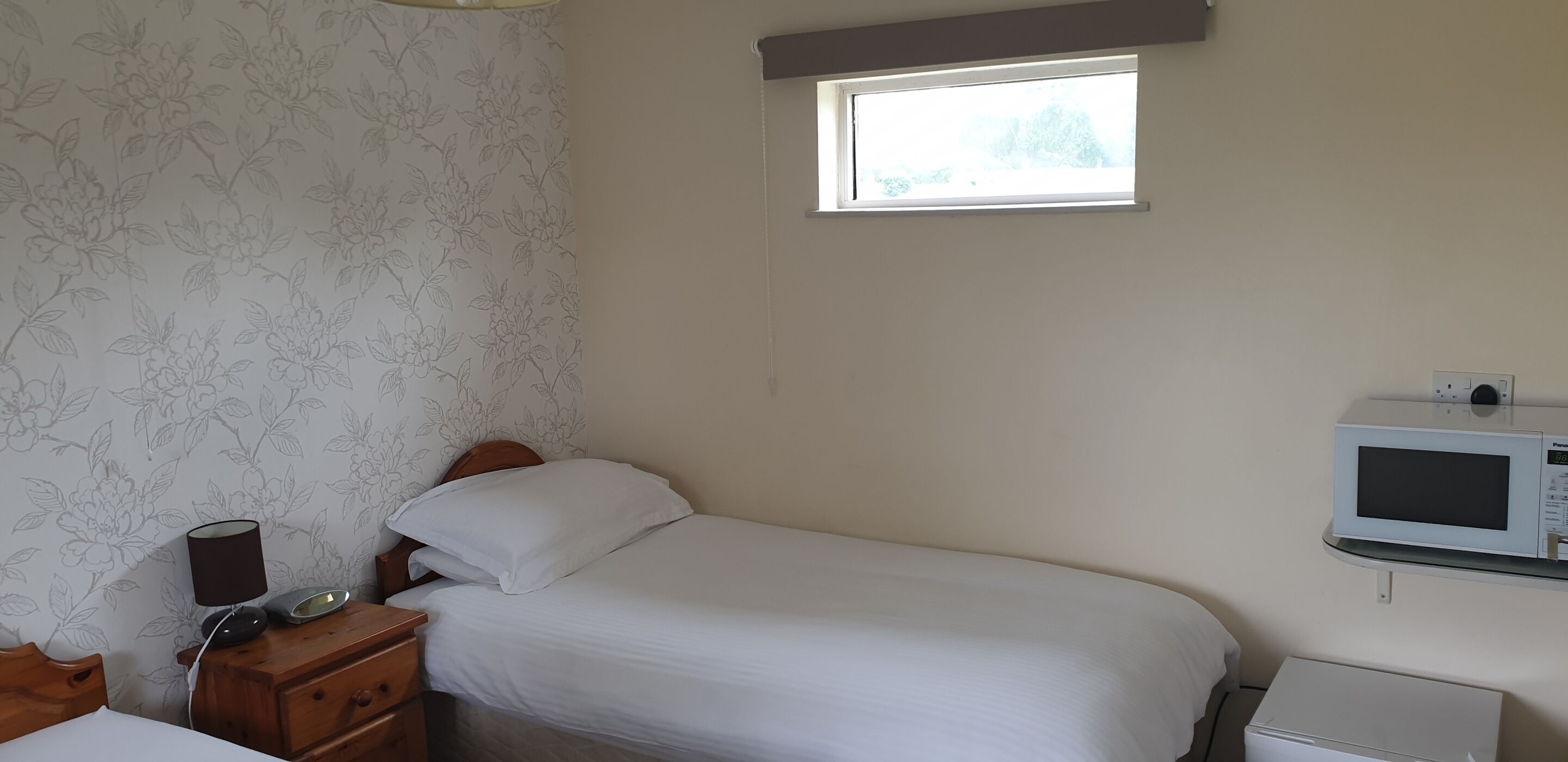 Een kleine, verzorgde slaapkamer met een eenpersoonsbed, een nachtkastje met lamp, een klein dressoir, een magnetron en een raam dat natuurlijk licht binnenlaat.
