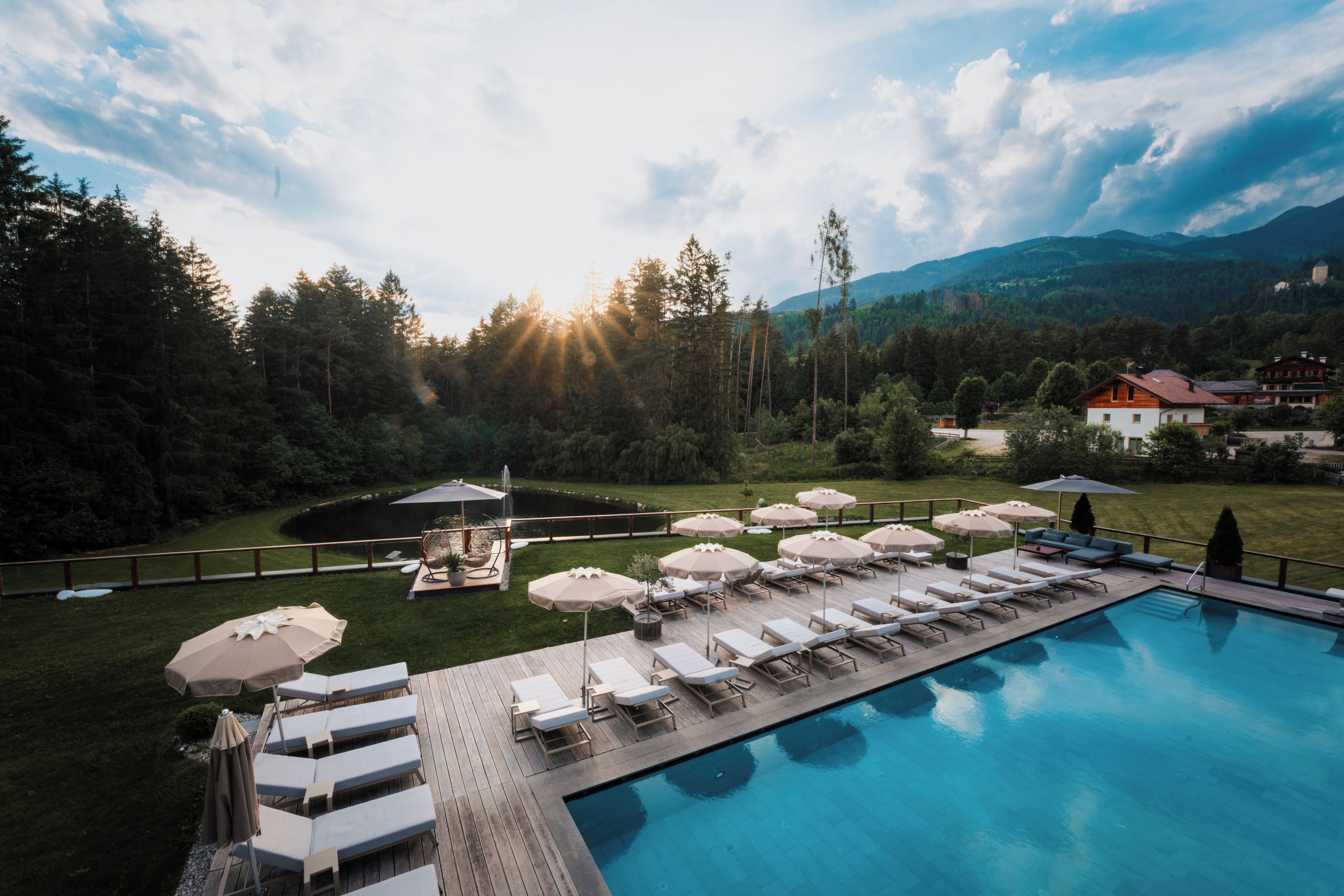 Un espace aménagé au bord de la piscine, avec des chaises longues et des parasols, entouré d’un paysage boisé et avec des montagnes en arrière-plan.