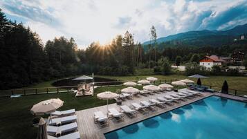 A poolside area with lounge chairs and umbrellas, surrounded by a forested landscape and mountains in the background.