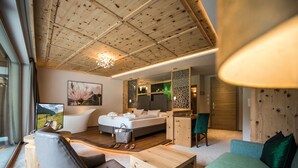 A modern hotel room with a wooden ceiling, a large bed, a sitting area with a sofa and armchair, a desk, and a view of the mountains. - Green Lake Hotel Weiher (Pfalzen)