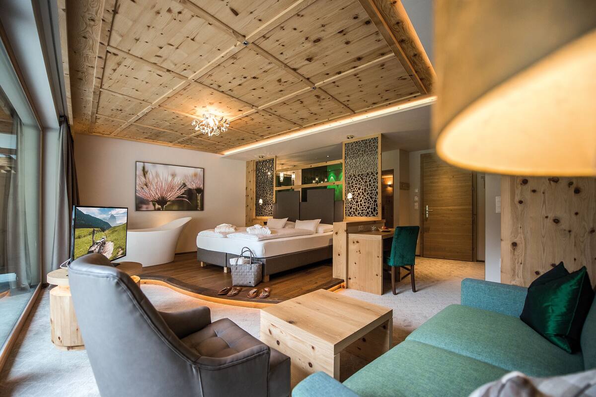 Habitación de hotel moderna con techo de madera, una cama grande, una zona de estar con sofá y sillón, un escritorio y vistas a las montañas.