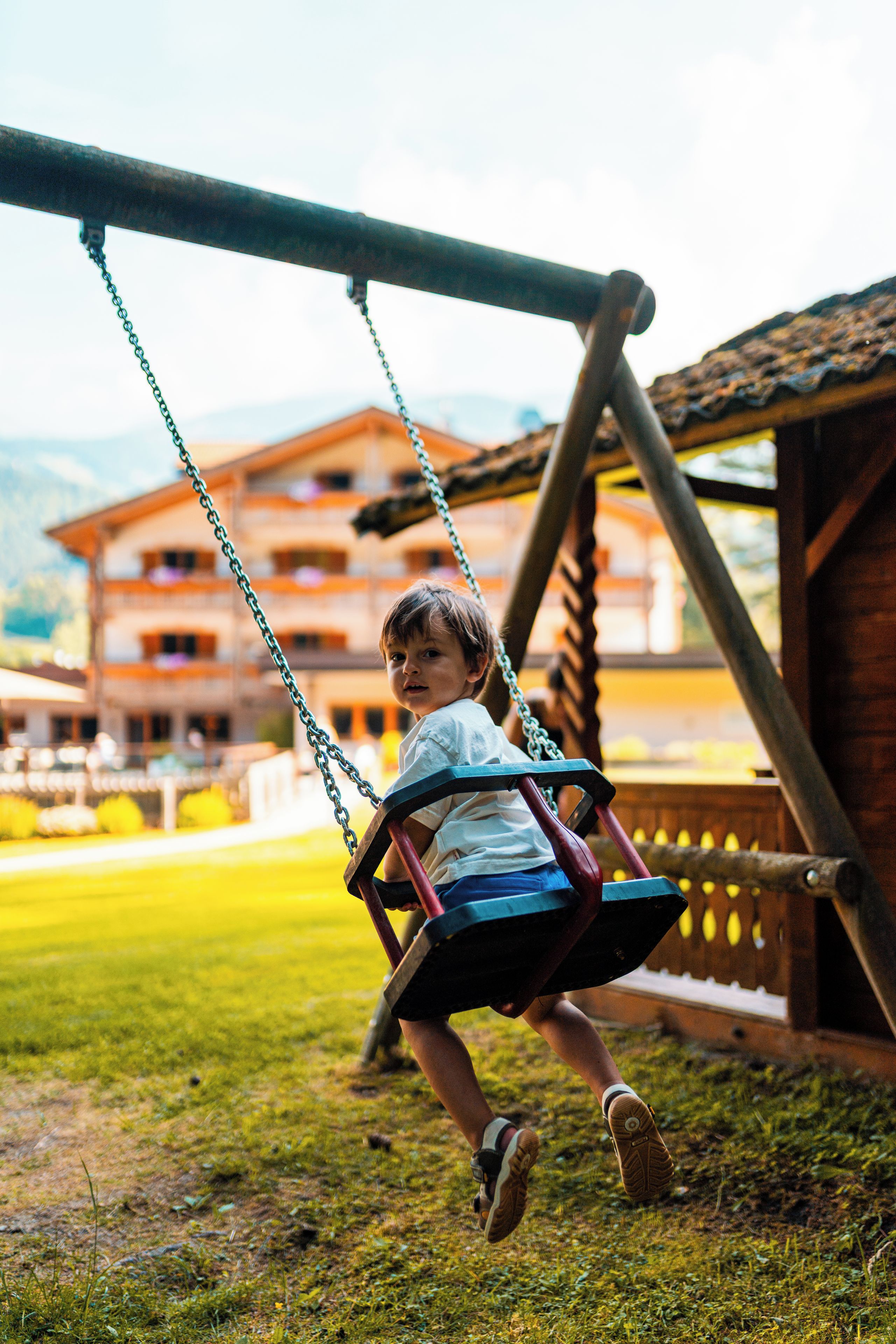 Un niño en un columpio en un área con césped, una estructura de madera y un edificio al fondo.
