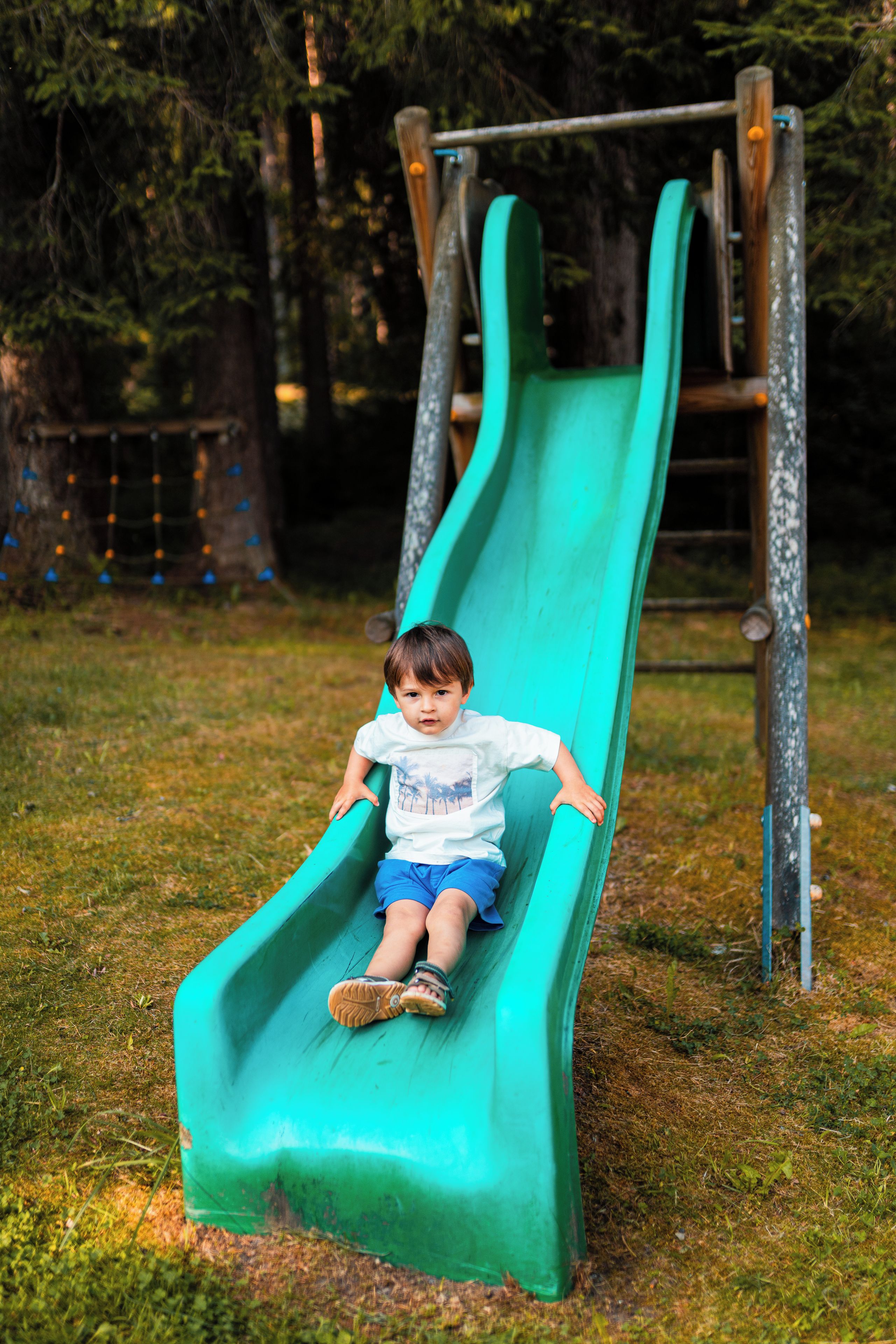 A child on a green slide in a park with trees and a playground structure in the background.