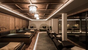A spa room with wooden paneling, multiple massage beds, a central seating area, and a chandelier. - Green Lake Hotel Weiher (Pfalzen)