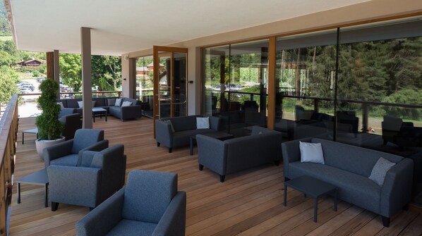 A modern outdoor lounge area with wooden flooring, grey sofas, and large windows offering views of a forested area.