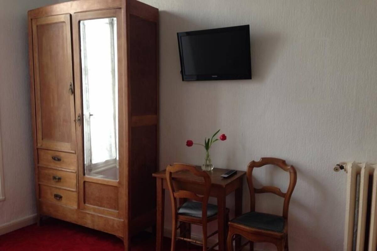 Una habitación con un armario de madera, un televisor montado en la pared, una mesita con sillas y un radiador.