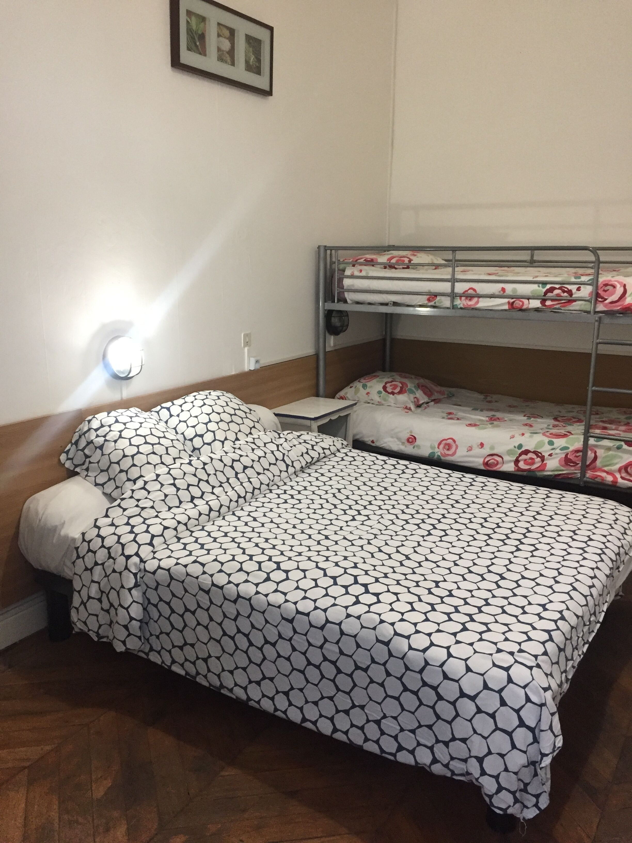 A single bed with a patterned bedspread, a bedside lamp, and a bunk bed with floral bedding in a room with wooden flooring.