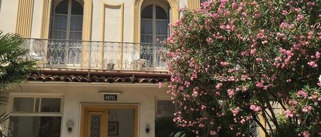 A hotel with ornate architectural details, a balcony, and a large flowering tree in front.